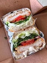 A close-up, high-quality shot of a chicken caesar sandwich served in a takeaway box. The image highlights the fresh ingredients, including the breaded chicken, crisp lettuce, tomato, and generous amount of dressing, all held within slices of focaccia.