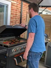 A photo of a man standing in a garden, cooking on a Blackstone griddle. The shot is taken from behind the subject, focusing on the griddle surface which contains indistinct, out-of-focus food items.