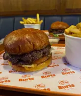 A close-up, eye-level shot of a double beef patty burger served on branded paper, with a cup of fries and a second burger visible in the blurred background, typical of a casual burger restaurant setting.