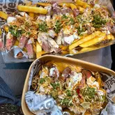 A top-down shot of two takeaway containers filled with steak and chips, presented on a lap inside a car. The lighting is bright and natural, highlighting the textures of the seared meat, crispy chips, and toppings.