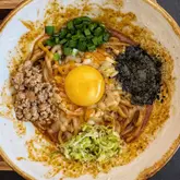 A top-down, high-resolution shot of a bowl of Japanese mazemen (stirred noodles). The dish is topped with a raw egg yolk, minced meat, chopped scallions, shredded seaweed, and melted cheese. The lighting is even and bright, highlighting the textures of the ingredients.