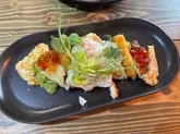 A casual, top-down photograph of a brunch dish featuring poached eggs, avocado, halloumi, and chili jam, served on a dark plate against a wooden table background.