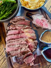 A high-angle, overhead shot of a premium gastropub meal featuring a sliced côte de boeuf steak, a cast-iron skillet of mac and cheese, broccolini, and two dipping sauces served on a wooden board.