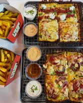 A high-quality, overhead flat-lay photograph of two Detroit-style pizzas and two portions of fries, presented on metal wire racks and branded takeaway boxes. The lighting is bright and even, effectively highlighting the textures of the crispy crusts, melted cheese, and various toppings.