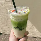 A bright, eye-level shot of a matcha latte in a clear takeaway cup, held by a hand against a neutral brick wall. The drink is topped with whipped cream and rainbow sprinkles, creating a vibrant and inviting aesthetic.