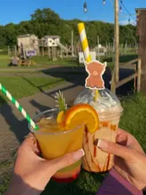 A bright, golden-hour shot of two hands holding a cocktail and a milkshake at Bluebell Dairy, with a playground in the background.