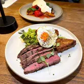A high-quality, well-lit food photograph featuring a brunch dish of flat iron steak, fried egg, smashed avocado, and a sweet potato hash brown, with a French toast dish visible in the background.
