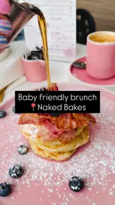 A short, high-quality close-up shot of syrup being poured over a stack of pancakes topped with bacon and blueberries at a cafe.
