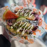 A close-up, top-down shot of a Mexican torta served in a branded paper-lined basket, held by a person. The image captures the messy, casual nature of the street food, highlighting the ingredients like red cabbage, cilantro, and lime.