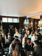 A candid, wide-angle shot capturing the busy, atmospheric interior of a pub. The image focuses on the social environment, with patrons seated at tables enjoying drinks and food, conveying a lively and welcoming vibe.