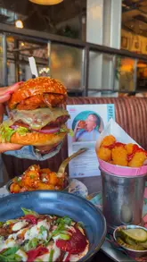 A promotional video for a collaboration menu between Poppy Cooks and Bill's restaurant. The video features close-up shots of a burger, loaded tater tots, a potato salad, and two cocktails, set to music with text overlays highlighting the items.