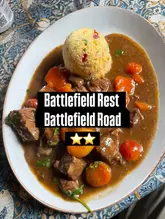 A top-down, amateur-style photograph of a lamb tagine dish served with a dome of couscous, presented on a patterned tablecloth. The image includes a text overlay identifying the venue and a two-star rating, consistent with a casual review format.