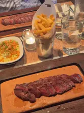 A high-angle, appetizing shot of a steak served on a wooden board, accompanied by a metal cup of fries and a small bowl of beef dripping popcorn, capturing the signature presentation of a Flat Iron restaurant.