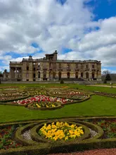 A high-quality, wide-angle landscape photograph of Witley Court and Gardens, featuring the historic ruined mansion set against a bright blue sky with fluffy clouds, framed by vibrant, well-manicured floral gardens in the foreground.