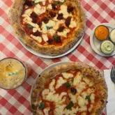 A top-down, flat-lay photograph of two pizzas served on a classic red and white checkered tablecloth, accompanied by a glass of drink and a side of dipping sauces.