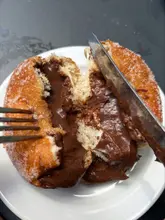 A close-up, top-down shot of a sugar-coated chocolate-filled donut being cut open with a knife and fork, revealing a rich, gooey chocolate center on a white plate.