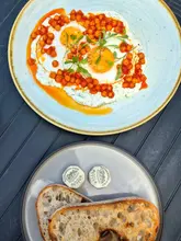 A bright, overhead food photography shot capturing a brunch spread. The main subject is a plate of Turkish eggs with chickpeas, accompanied by a separate plate of sourdough toast and butter. The natural lighting enhances the vibrant colours of the dish, and the composition is clean and well-balanced.
