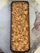 A top-down shot of a homemade gluten-free Bakewell tart, featuring a frangipane filling topped with slivered almonds, presented in a rectangular tart tin on a textured surface.