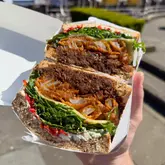 A close-up, handheld shot of a 'Laughing Cow' sandwich, featuring black treacle beef, onion rings, cream cheese, and chillies, presented in a takeaway box.