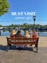 A bright, sunny, and inviting lifestyle shot featuring four people sitting on a bench overlooking a marina. The composition is symmetrical and clean, effectively showcasing the scenic, relaxing atmosphere of the location. The text overlay clearly communicates the value proposition and the location's accessibility, making it an effective piece of travel and lifestyle marketing content.