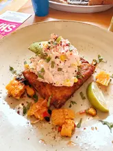 A top-down, slightly angled shot of a salmon hash dish served at a cafe, featuring a hash brown topped with a salmon and corn mixture, garnished with herbs and a lime wedge.