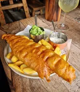 A high-quality, close-up shot of a classic fish and chips meal served in a pub setting. The focus is sharp on the golden, crispy batter of the fish, with sides of mushy peas and tartare sauce presented in small metal pots. The rustic wooden table and branded paper add to the authentic pub atmosphere.