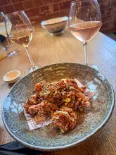 A high-quality, well-composed shot of a Japanese fried chicken dish served in a branded bowl, accompanied by two glasses of wine. The lighting is natural and flattering, highlighting the texture of the food, while the shallow depth of field creates a professional, inviting atmosphere.