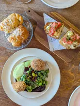 A high-quality, overhead flat-lay photograph showcasing a selection of dishes from a tasting menu. The image features a plate of croquettes with a vibrant green pea and broad bean salsa, a wooden board with tomato-topped bread, and a glass plate with pastries. The lighting is bright and even, highlighting the textures and colours of the food, while the simple wooden table background keeps the focus entirely on the presentation.