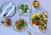 A bright, well-composed overhead flat-lay photograph of a lunch spread at a restaurant. The image features a variety of small plates including labneh, burrata, salt and pepper squid, and fries, arranged neatly on a table. The natural lighting is consistent and flattering, highlighting the textures and colors of the food.
