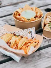 A high-angle, natural light shot of a tray of tacos from a street food market, set on a wooden picnic table. The primary subject is the tacos in the foreground, with a bowl of tortilla chips and a container of dumplings visible in the background.
