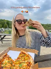 A casual, user-generated style shot of a person eating a pizza slice outdoors, likely at a pop-up or casual venue. The image captures the enjoyment of the food but suffers from a cluttered background and tight framing.
