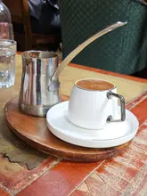 A close-up shot of a traditional Turkish coffee set, featuring a small white cup and a metal cezve on a wooden serving board, set against a rustic table surface.