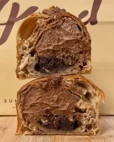 A high-quality, close-up cross-section shot of a chocolate-filled pastry, likely a cruffin, stacked on a wooden surface. The image effectively showcases the lamination of the pastry dough and the generous, rich chocolate filling, with the bakery's branded paper bag providing context in the background.