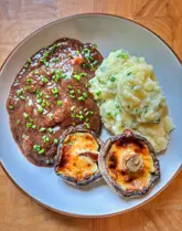 A top-down shot of a home-cooked meal featuring beef bourguignon, creamy mashed potatoes, and two stuffed mushrooms, served on a white plate on a wooden surface.