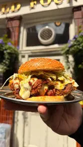Here are a few options for your SEO alt text (under 125 characters):

**Option 1 (Descriptive):**
Chef James Cochran preparing a Bird and Boujee fried chicken burger at The Old Fountain pub in London.

**Option 2 (Focus on food):**
Close-up of a gourmet fried chicken burger with truffle gravy and blue cheese from Around The Cluck, London.

**Option 3 (Action-oriented):**
Chef James Cochran serving a luxury fried chicken burger to a customer at The Old Fountain pub.