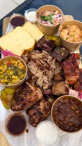 A promotional video for a BBQ takeaway pop-up in South Wales, featuring a large sharing platter with various smoked meats, sides, and a banana pudding dessert. The video uses close-up shots to highlight the texture and quality of the food.