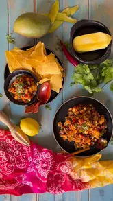 A top-down recipe tutorial demonstrating the preparation of a mango salsa, featuring fresh ingredients like mango, cilantro, and chili, served with tortilla chips.