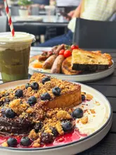A high-quality, well-composed food photography shot featuring a plate of berry-topped French toast in the foreground, with a full English breakfast and a matcha latte visible in the background, set on an outdoor table.