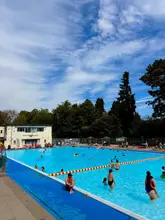 A bright, wide-angle shot of an outdoor lido on a sunny day, capturing the scale of the pool and the surrounding environment. The image serves as a functional venue tour, showcasing the facility's atmosphere and popularity.