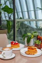 A high-quality, well-lit photograph of two stacks of pancakes at Darwin Brasserie in the Sky Garden, London. One stack features berries and cream, while the other has caramelised banana, Nutella, and hazelnuts, accompanied by a latte.
