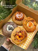 A close-up, top-down shot of a box containing four distinct, creatively topped Korean-style cookies, held in a hand against a blurred outdoor background.