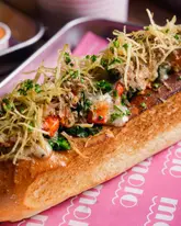 A high-quality, close-up food photograph of a lobster roll topped with crispy shoestring potatoes and fresh chives, served on branded pink paper. The lighting is professional and highlights the textures of the seafood and the toasted bun.