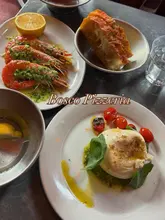 A casual, overhead flat-lay shot of a lunch spread at Bosco Pizzeria, featuring burrata with roasted tomatoes and pesto, focaccia, and garlic prawns. The image captures a standard restaurant dining experience.