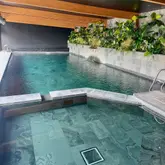 A high-quality, well-composed photograph showcasing an indoor swimming pool at a luxury accommodation venue. The image highlights the serene atmosphere, featuring clean lines, natural stone tiling, and a lush green wall, effectively communicating a relaxing and premium experience.