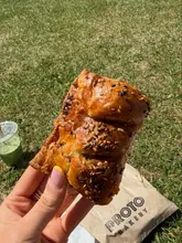 A close-up, handheld shot of a sausage roll, likely taken outdoors on grass, with a bakery bag and a green drink visible in the background.