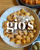 A high-angle, top-down food photography shot of a bowl of rigatoni pasta with a rich, reddish sauce, topped with grated cheese and herbs. A small bowl of olives is visible in the bottom right corner, and another pasta dish is partially visible at the top. The image features a prominent white text overlay branding the venue as 'gio's' with the location 'Slateford Road'.