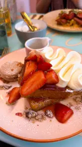 A short, aesthetic reel showcasing a brunch visit to 'Let Them See Cake'. The video features close-up shots of French toast being drizzled with chocolate, a savoury brunch plate, a selection of cakes in a display cabinet, and a latte, ending with a shot of the shop exterior.