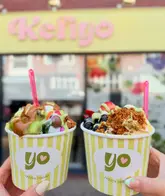 A bright, well-composed shot of two cups of frozen yogurt held in front of the 'Kefiyo' storefront, showcasing generous toppings and sauces.