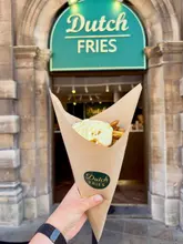 A first-person perspective shot of a paper cone filled with Dutch-style fries topped with mayonnaise, held in front of the 'Dutch Fries' venue entrance. The lighting is bright and natural, highlighting the texture of the fries and the creamy sauce.