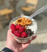 A close-up, first-person perspective shot of a yogurt bowl held in a hand, featuring toppings like raspberries, peanut butter, coconut, and chocolate chips.